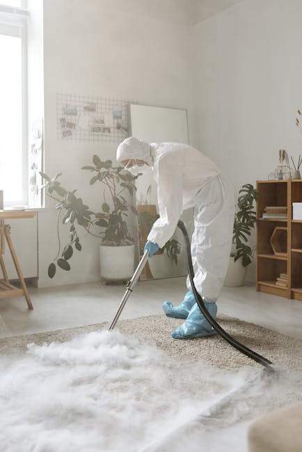 A professional cleaner dressed in a white protective suit, face mask, and blue gloves is using a vacuum cleaner to perform deep carpet cleaning in a bright, contemporary living room. The room features a beige area rug on a light-colored wooden floor, with sunlight streaming through a window on the left, illuminating potted plants and minimalist wooden furniture. There are decorative items on a wooden shelving unit and a whiteboard or corkboard on the wall. The scene highlights thorough surface cleaning and the emphasis on hygiene, consistent with services offered by Deep Cleaning Kensington in South Kensington near Gloucester Road.