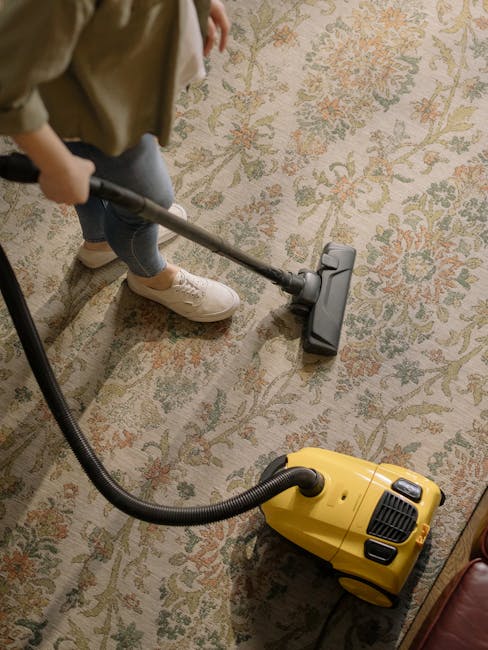 A child wearing beige shoes, jeans, and a khaki jacket is vacuuming a patterned, traditional-style rug with a yellow and black vacuum cleaner. The rug features intricate floral motifs in muted tones of green, orange, and beige, covering a large area of the wooden floor. The vacuum cleaner's hose is connected to a handheld attachment, which the child is holding to clean the rug thoroughly. Natural light illuminates the scene, highlighting the surface cleanliness and the detailed pattern of the rug, with the vacuuming process illustrating aspects of surface cleaning and deep cleaning within a residential setting. Deep Cleaning Kensington specializes in professional domestic cleaning services, including carpet and rug cleaning to maintain hygiene and enhance appearance.