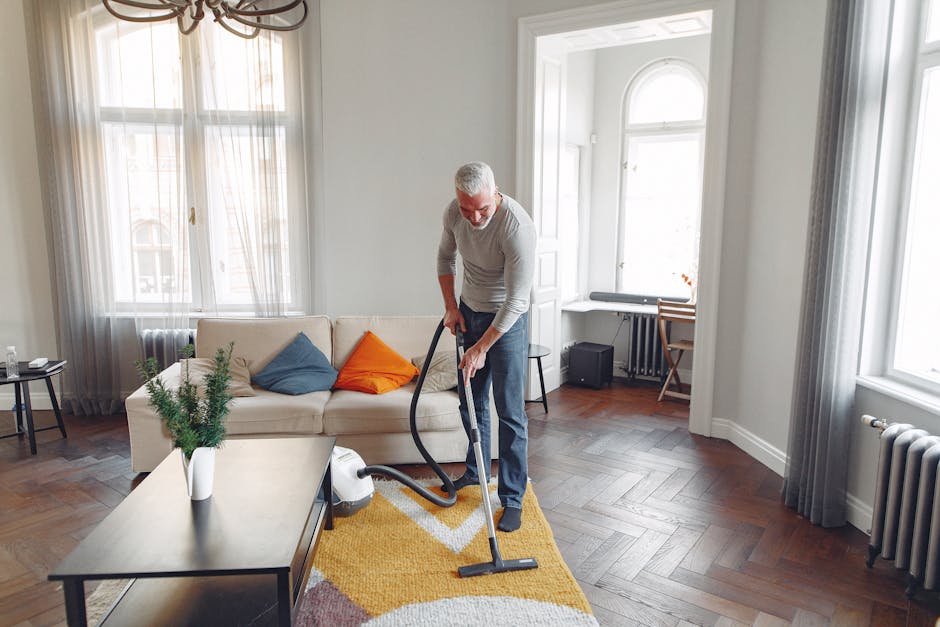 A man with gray hair and glasses is vacuuming a living room with a beige sofa, decorated with orange and blue cushions, a yellow patterned rug, and a white coffee table with a small green potted plant. The room features large windows with sheer curtains, white walls, and wooden flooring styled in a herringbone pattern. There is a desk and chair near the window, a radiator, and a small black speaker on the floor, all illuminated by natural light. Deep Cleaning Kensington, a domestic cleaning service, is demonstrating proper surface cleaning and deep cleaning techniques in a well-lit, tidy living space to avoid common carpet cleaning mistakes in Kensington flats.