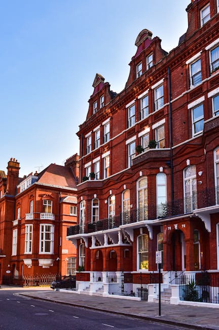 Exterior view of a red brick Victorian flat building near High Street Kensington, featuring large sash windows with white frames, decorative stone details, and black wrought-iron balcony railings. The building's ground floor has a white entrance porch with ornate wooden supports and steps leading up to a wooden door. The surrounding street is paved and clean, with minimal road markings and a clear blue sky overhead. The scene emphasizes the historic architecture, illuminated by natural daylight, suggesting a well-maintained residential property, indicative of professional deep cleaning and maintenance services offered by Deep Cleaning Kensington for Victorian flats near High Street Kensington.