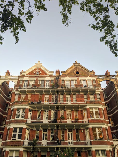 A person walking on a residential sidewalk near Victorian-style red-brick flats close to High Street Kensington, with two dogs on leashes. The sidewalk is clean and well-maintained, with black iron fencing along the street and some greenery from nearby trees. The buildings feature ornate architectural details, bay windows, and decorative columns, illuminated by natural light during daytime. The scene highlights the urban residential environment requiring routine cleaning and upkeep, as provided by Deep Cleaning Kensington, a professional cleaning company specializing in deep cleaning and surface sanitisation for such Victorian flats.