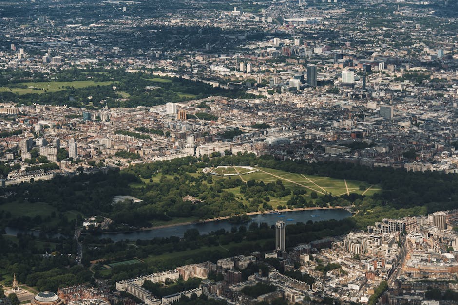 Aerial view of Hyde Park in Kensington, London, showing expansive green lawns, walking paths, and a pond, surrounded by urban buildings and streets. The park is lush with trees and appears well-maintained, providing a contrast to the dense cityscape. Bright sunlight highlights the vibrant greenery and reflective surfaces, with a mixture of residential and commercial structures in the background. This setting exemplifies residential and commercial urban landscaping and the importance of outdoor space maintenance, aligning with Deep Cleaning Kensington's focus on surface cleaning and deep cleaning services for homes in Kensington area.