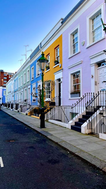 Image of a row of colorful terraced houses on a street in Kensington, including pastel blue, yellow, and lavender buildings. The houses feature white-framed windows and black iron railings along small front steps. A traditional black street lamp is positioned on the sidewalk, which is paved with grey slabs. The scene is well-lit with bright daylight, highlighting the clean and well-maintained appearance of the exterior facades. This vibrant street scene exemplifies high-quality property presentation, aligned with residential deep cleaning and maintenance services offered by Deep Cleaning Kensington, as featured on their page about oven cleaning for homes in Kensington.